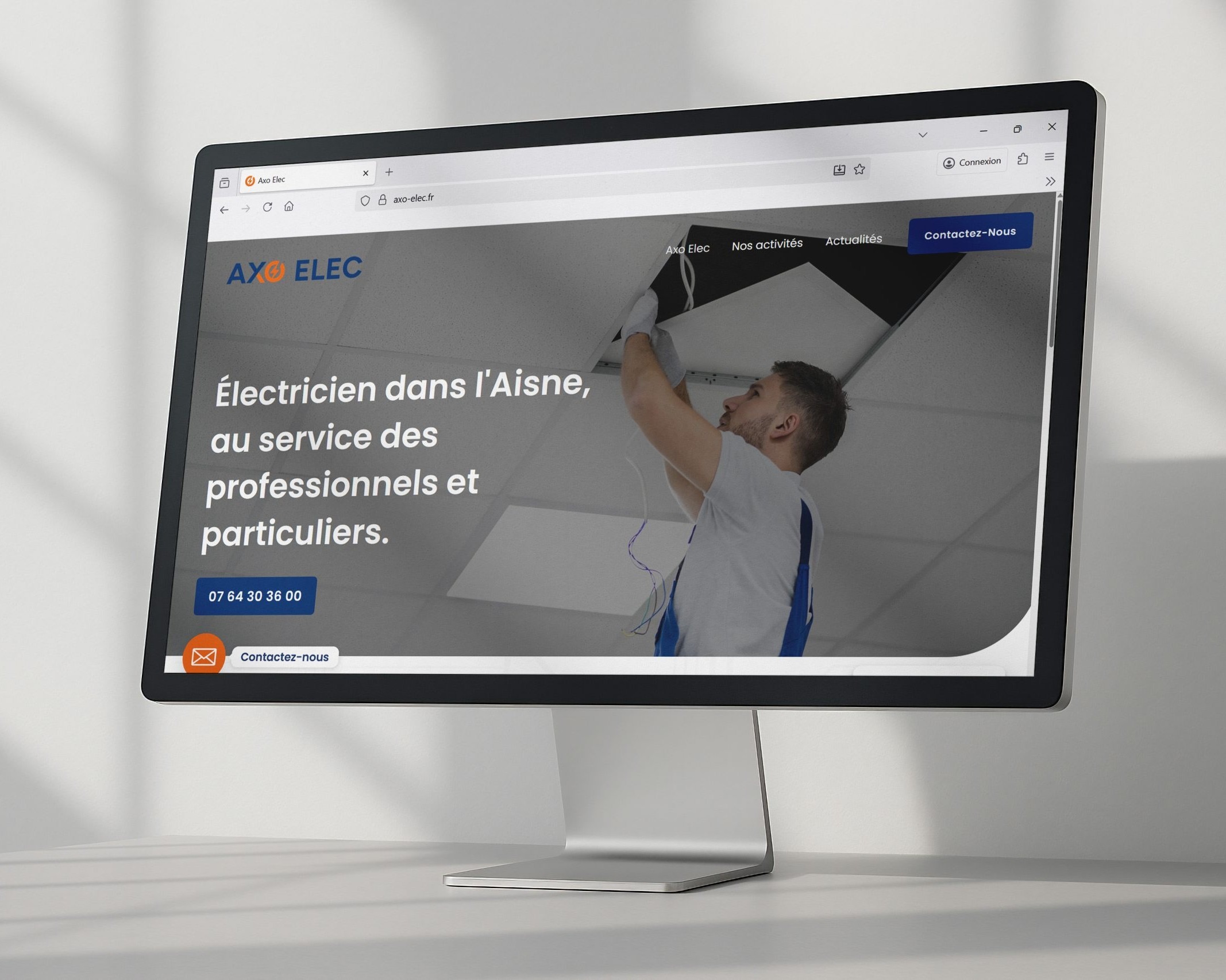 Screenshot of Axo Elec website on a tilted monitor, showing a technician wiring a ceiling and a French headline about electricians for professionals and individuals.